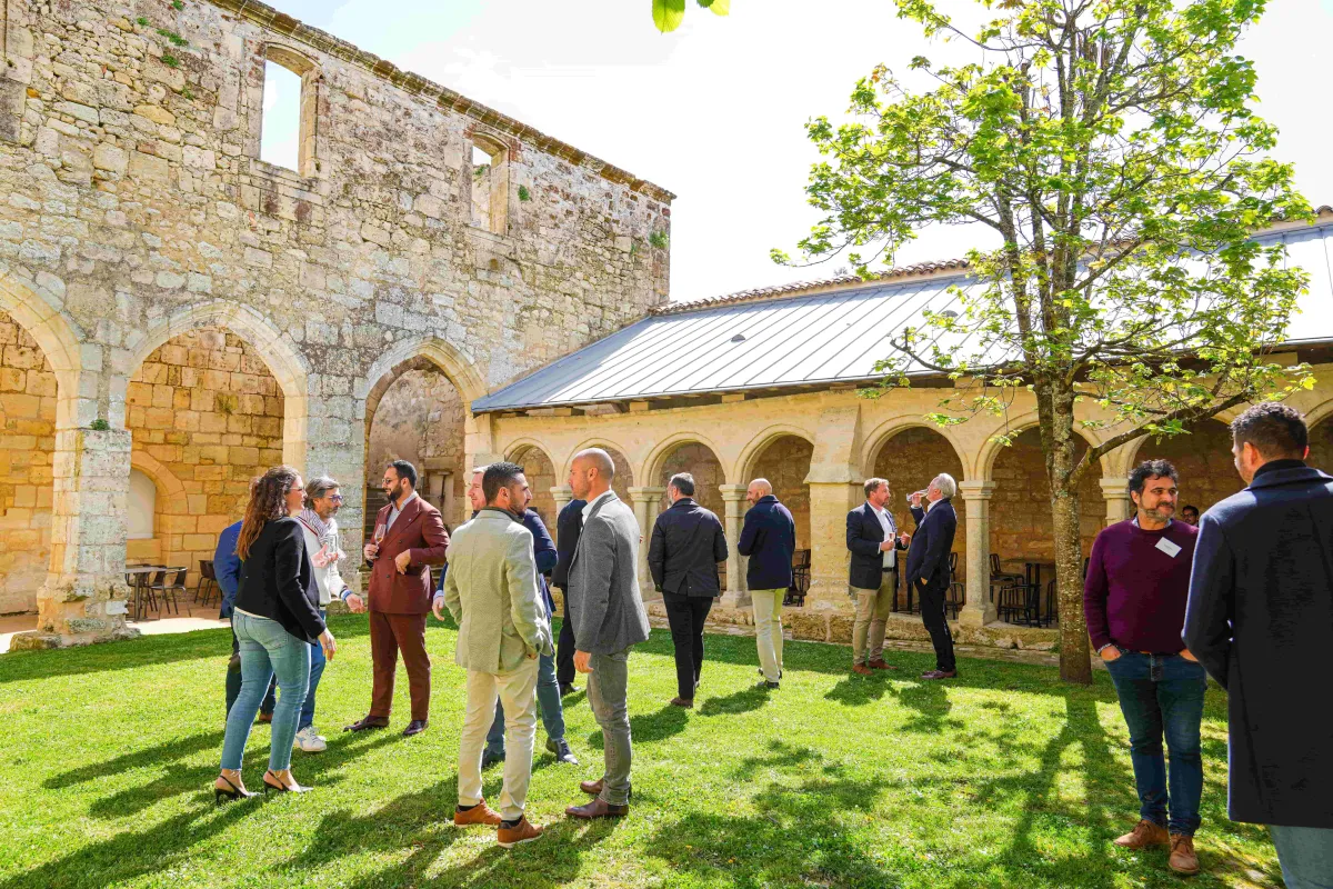 Groupe de professionnels en réseau dans la cour d'un monastère historique en pierre avec des cloîtres voûtés, pelouse verte et architecture médiévale lors d'un séminaire d'entreprise à Saint-Emilion.