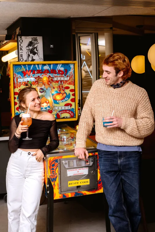 Deux personnes savourant des boissons à côté d'une machine de jeu d'arcade colorée et vintage dans un lieu moderne aux ambiances chaleureuses