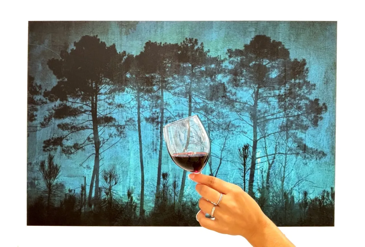 Main tenant un verre de vin rouge devant un paysage forestier avec de grands pins sous un ciel turquoise