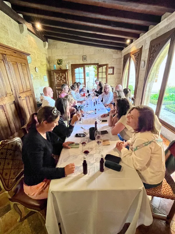 Un groupe de personnes assises autour d'une longue table draée de blanc lors d'un événement de repas formel dans une salle aux murs en pierre avec poutres en bois apparentes et grandes fenêtres donnant sur le paysage verdoyant.