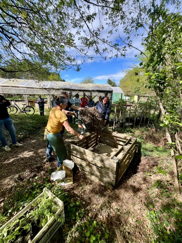 Groupe de personnes compostant des déchets organiques dans des bacs en bois lors d'un atelier de jardinage communautaire en plein air par une journée ensoleillée.