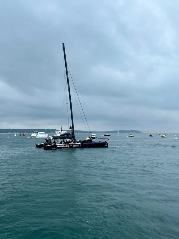 Un catamaran à voile noir avec un mât élancé mouillé dans une eau turquoise calme, entouré d'autres bateaux dans une baie côtière sous un ciel nuageux.