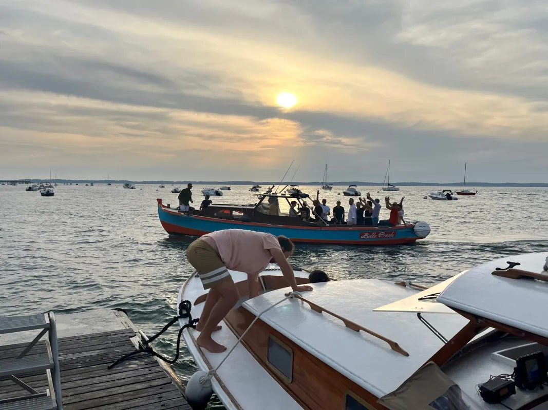 Un bateau de visite touristique bleu et rouge rempli de passagers sur des eaux calmes au coucher de soleil, avec des voiliers amarrés et de petits bateaux visibles en arrière-plan près d'une jetée en bois.