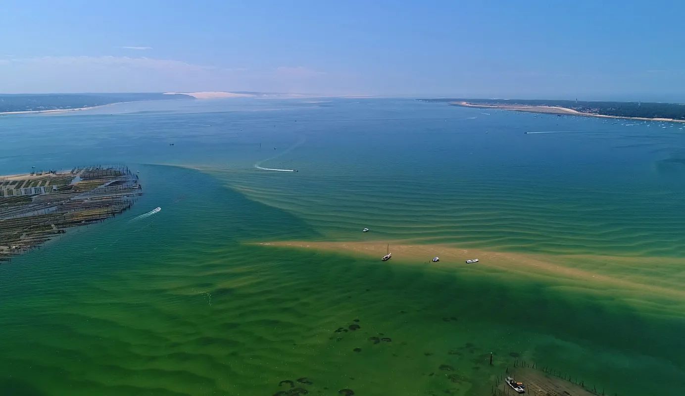 Vue aérienne du Bassin d'Arcachon montrant des eaux turquoises peu profondes, un grand navire de croisière amarré à gauche, des voiliers dispersés et des plages de sable entourées de forêts de pins.