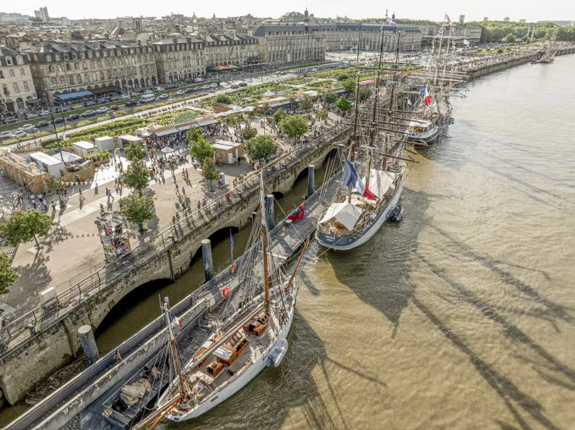 Vue aérienne du quai de Bordeaux avec des voiliers historiques amarrés, une place publique avec espaces verts et visiteurs, des immeubles haussmanniens, et la Garonne.