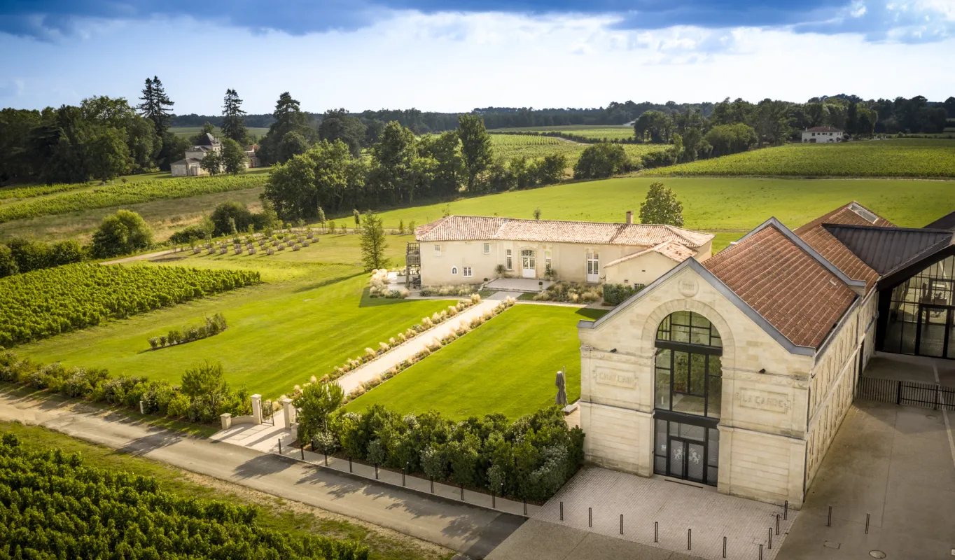 Vue aérienne du Château La Garde à Bordeaux, montrant un bâtiment moderne en pierre avec baies vitrées cintrées, jardins aménagés et vignobles étendus entourés de champs verts et d'arbres sous un ciel bleu.