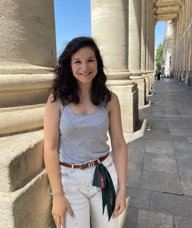 Jeune femme souriante devant les colonnes néoclassiques majestueuses de Bordeaux, vêtue d'un débardeur bleu clair et d'un jean blanc avec ceinture marron