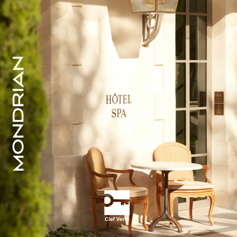 Entrée du spa de l'hôtel Mondrian Bordeaux, avec des chaises en bois élégantes et une petite table ronde dans une cour ensoleillée aux murs en pierre crème et lierre grimpant.