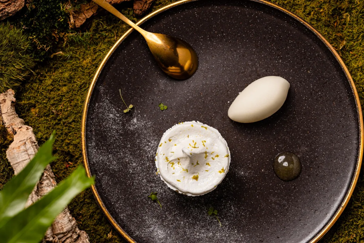 Une assiette noire à bordure dorée sur une surface couverte de mousse, garnie d'une mousse de fromage blanc parsemée de pistaches, d'un élément blanc en forme d'œuf, une cuillère dorée et une sphère translucide, entourés d'éléments naturels.