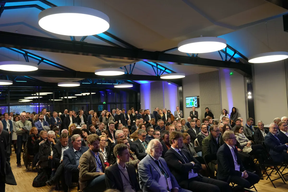Large audience seated and standing in a modern conference hall with blue ambient lighting and round ceiling lights, attendees wearing business attire with lanyards, watching a presentation on a screen.