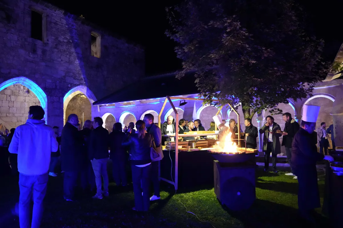 Soirée de gala dans un monument historique en pierre avec des convives rassemblés autour d'un foyer central, éclairés par des lumières bleues et chaudes, à Saint-Emilion, région de Bordeaux.