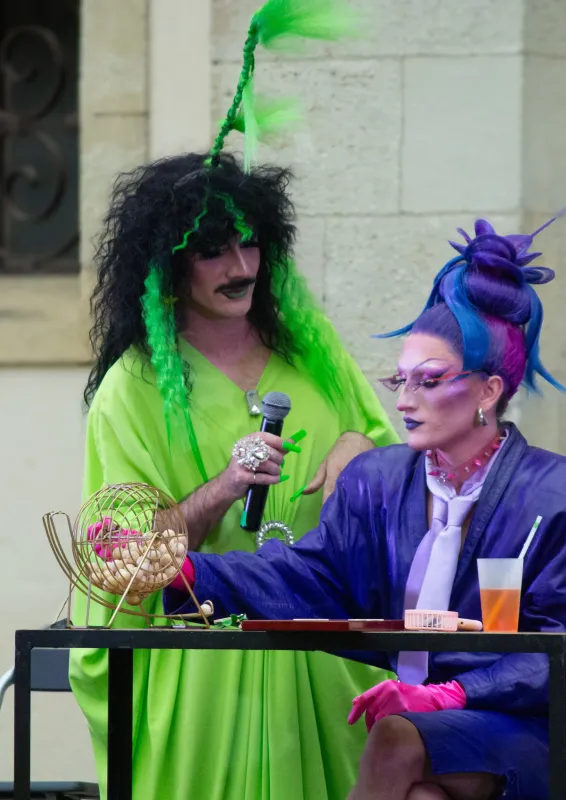 Deux performeurs lors d'un événement à Bordeaux : l'un vêtu d'une tenue vert fluo avec des cheveux noirs et des guirlandes vertes, tenant un microphone, à côté d'un autre performeur en tenue violette avec des cheveux bleu et violet hérissés, des lunettes 