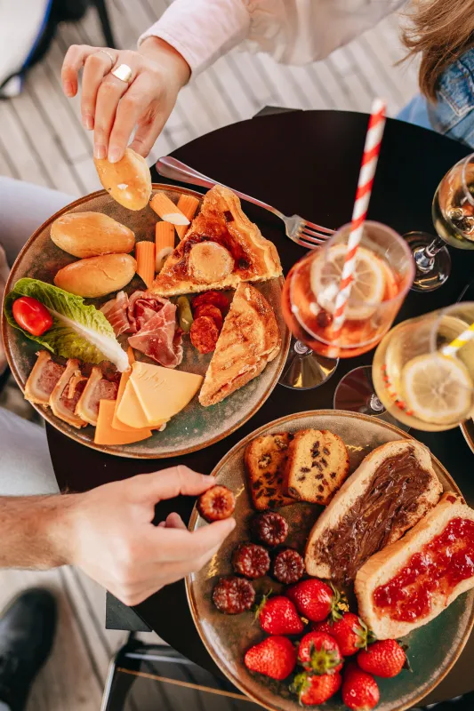 Deux personnes dégustant un brunch gastronomique avec charcuterie, viennoiseries, fruits frais et vin rosé sur une table noire lors d'une croisière sur la Garonne à Bordeaux
