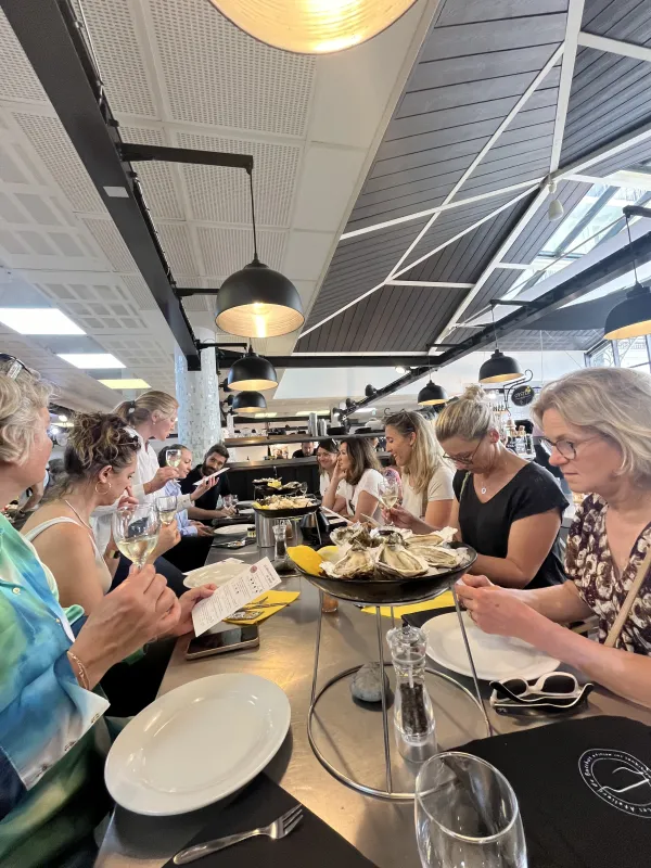 Groupe de personnes dégustant des mets et du vin dans un lieu de restauration moderne avec cuisine ouverte, plafond industriel et luminaires suspendus.