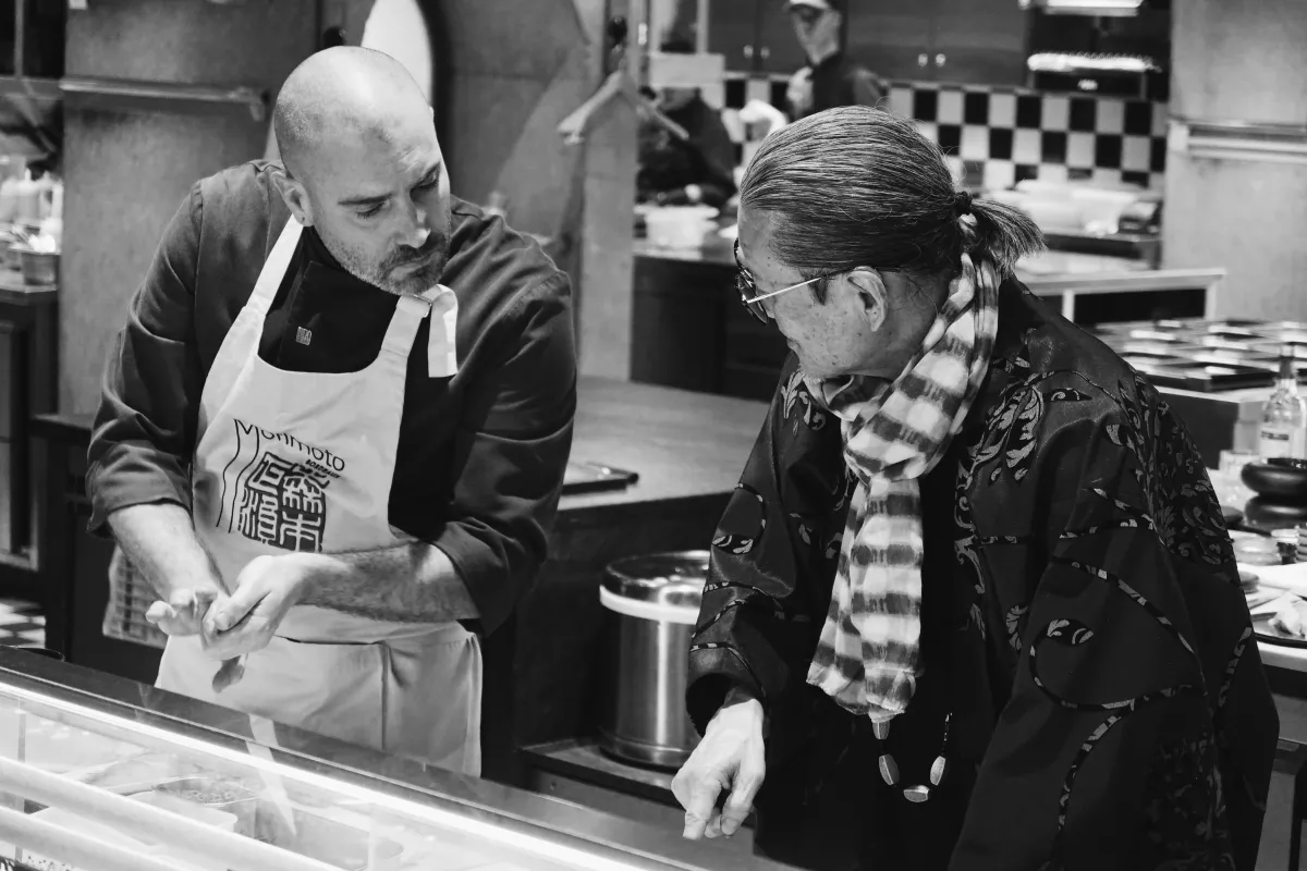 Un chef portant un tablier blanc avec des caractères asiatiques démontre une technique culinaire à une femme âgée portant des lunettes dans une cuisine professionnelle du Mondrian Bordeaux Les Carmes.