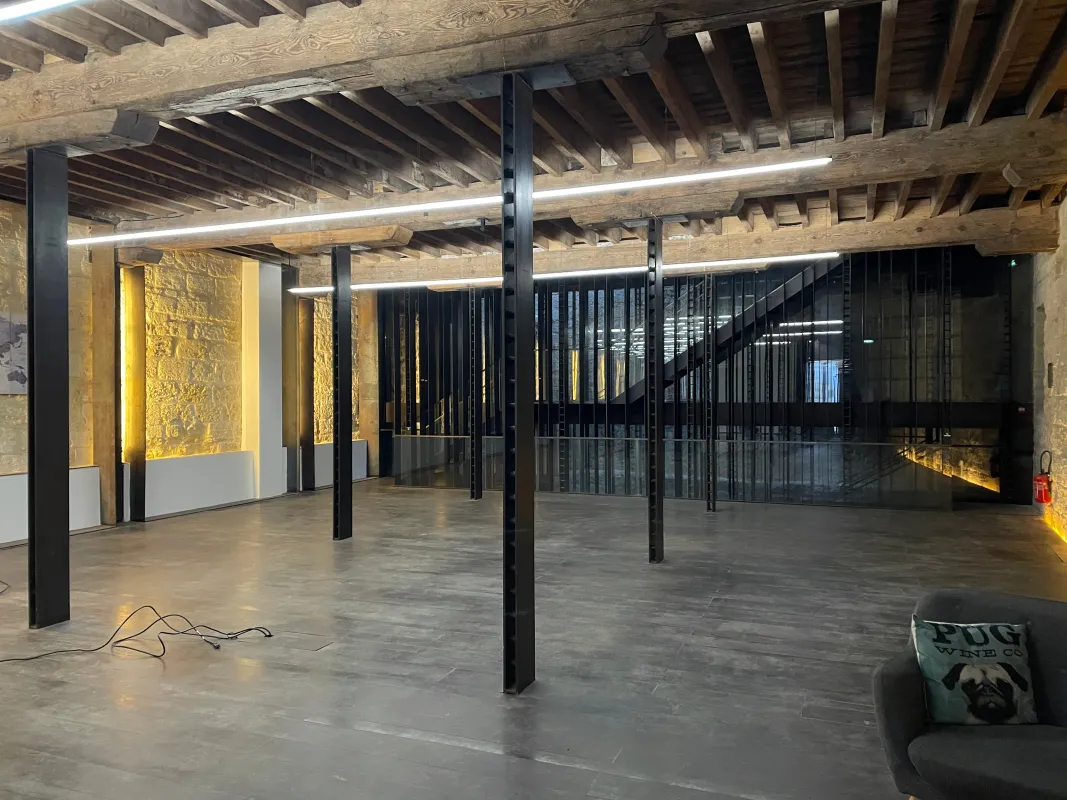 Espace loft industriel moderne avec poutres apparentes en bois, sol en béton, colonnes et garde-corps en acier noir, murs de brique éclairés en jaune, et luminaires contemporains à Bordeaux.