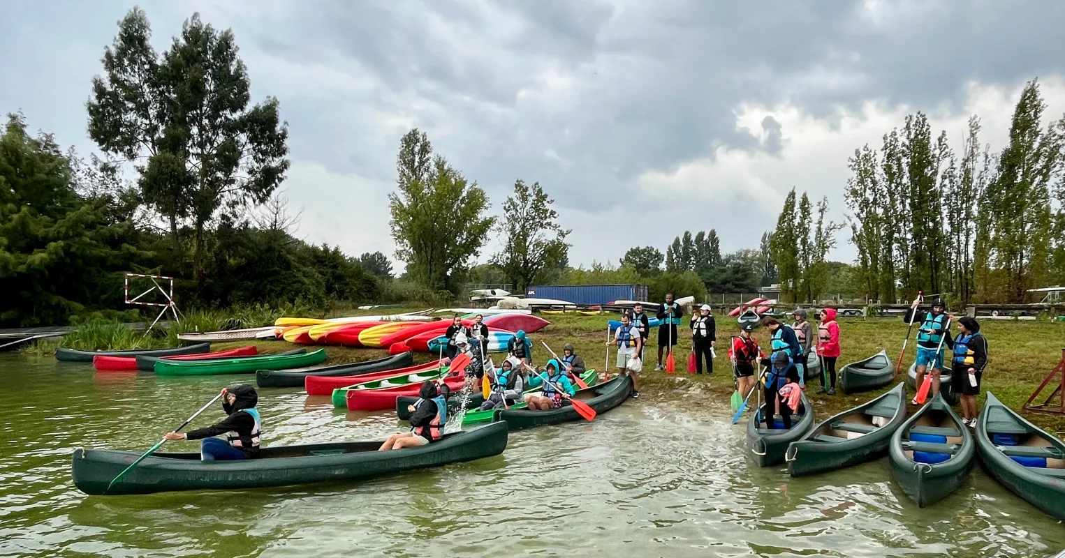 Un groupe de participants rassemblés dans un centre de sports nautiques avec plusieurs canoës et kayaks alignés sur la berge et dans l'eau, arbres et ciel nuageux en arrière-plan.