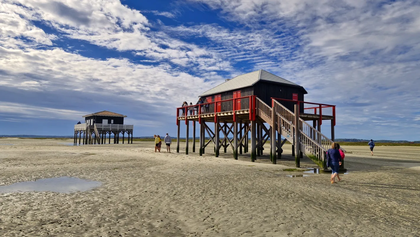 Maisons colorées sur pilotis sur une plage de sable avec des visiteurs explorant le littoral spacieux sous un ciel bleu