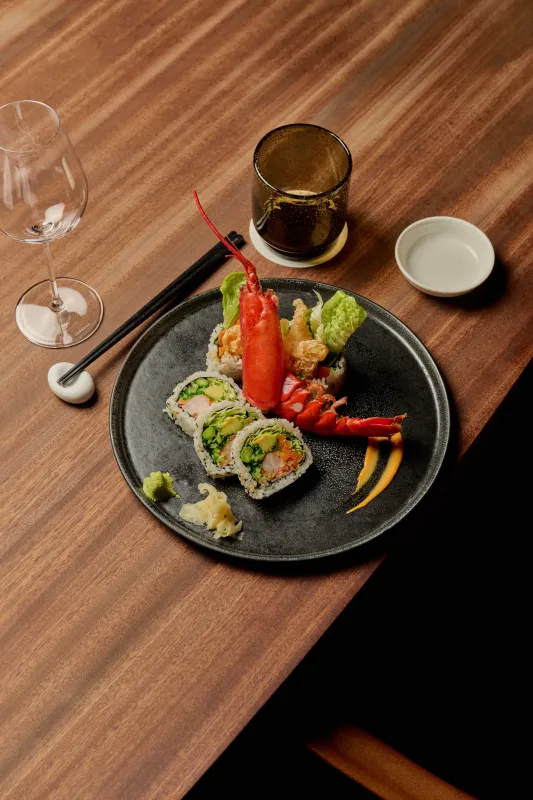 Assiette élégante de sushi avec des rouleaux de nigiri, des légumes frais et une garniture de poivron rouge sur une assiette grise foncée, accompagnée de vin blanc et de baguettes sur une table en bois