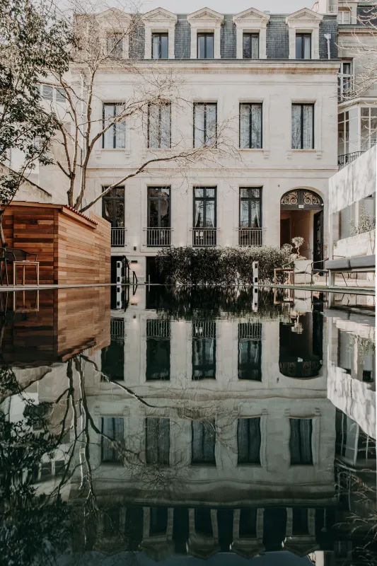 Un immeuble de style haussmannien se reflétant dans l'eau immobile, avec une structure moderne en bois sur la gauche, des arbres nus et des fenêtres symétriques dans une cour bordelaise.