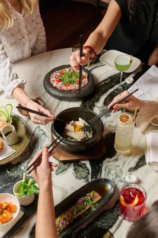 Deux personnes partageant un repas gastronomique autour d'une table avec une cuisine asiatique raffinée, comprenant des plats dans des bols noirs, des ingrédients frais et des boissons colorées sur une surface en marbre.