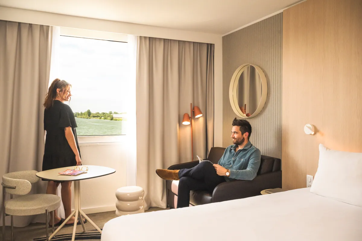 Chambre d'hôtel moderne du Novotel Bordeaux Lac avec deux clients, l'un debout près de la fenêtre donnant sur le lac et l'autre assis sur le canapé, avec mobilier contemporain et éclairage chaleureux