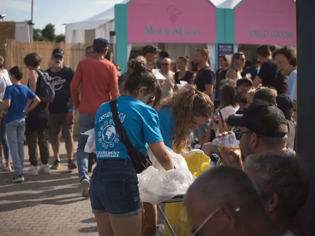 Un événement extérieur bondé à Bordeaux avec une bénévole en chemise bleue distribuant des articles aux visiteurs près de pavillons de marque roses et teals.