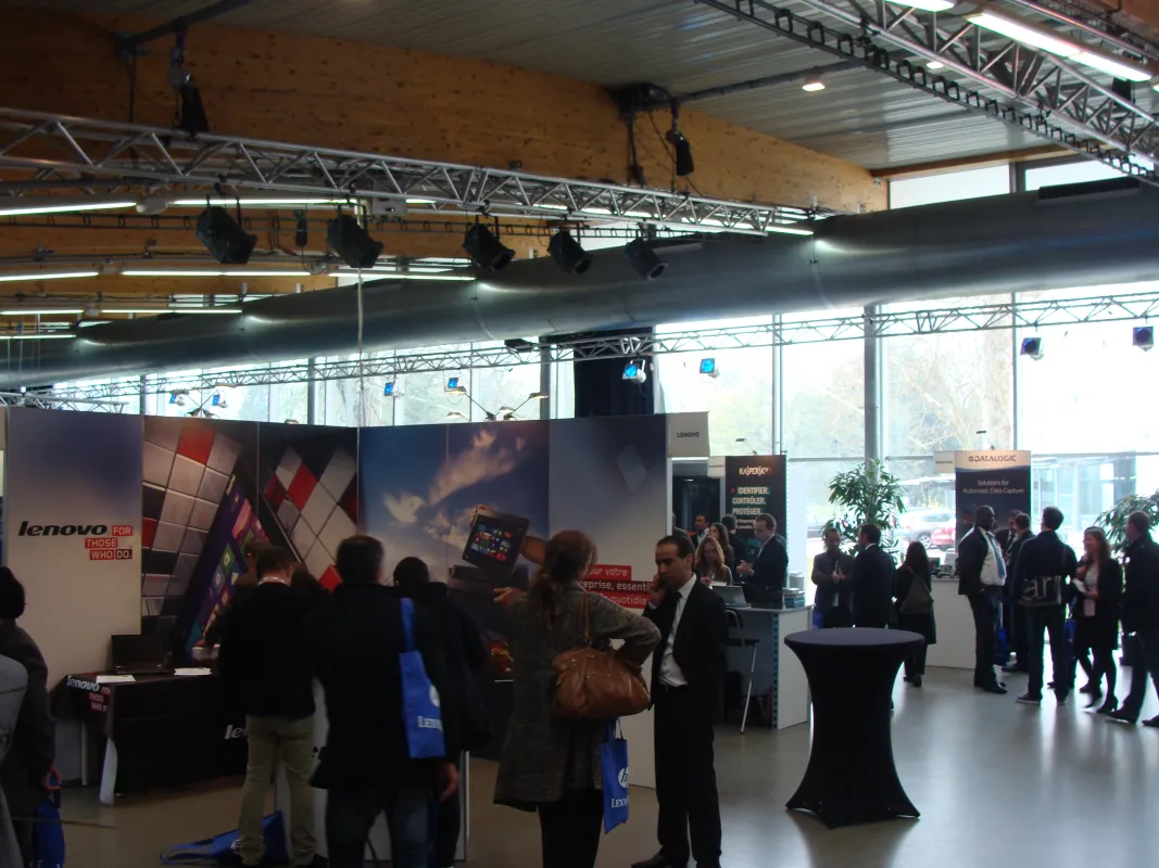 Salon professionnel intérieur avec stands d'exposition Lenovo, visiteurs en tenue professionnelle, et infrastructure moderne avec charpentes métalliques et panneaux d'affichage grand format.
