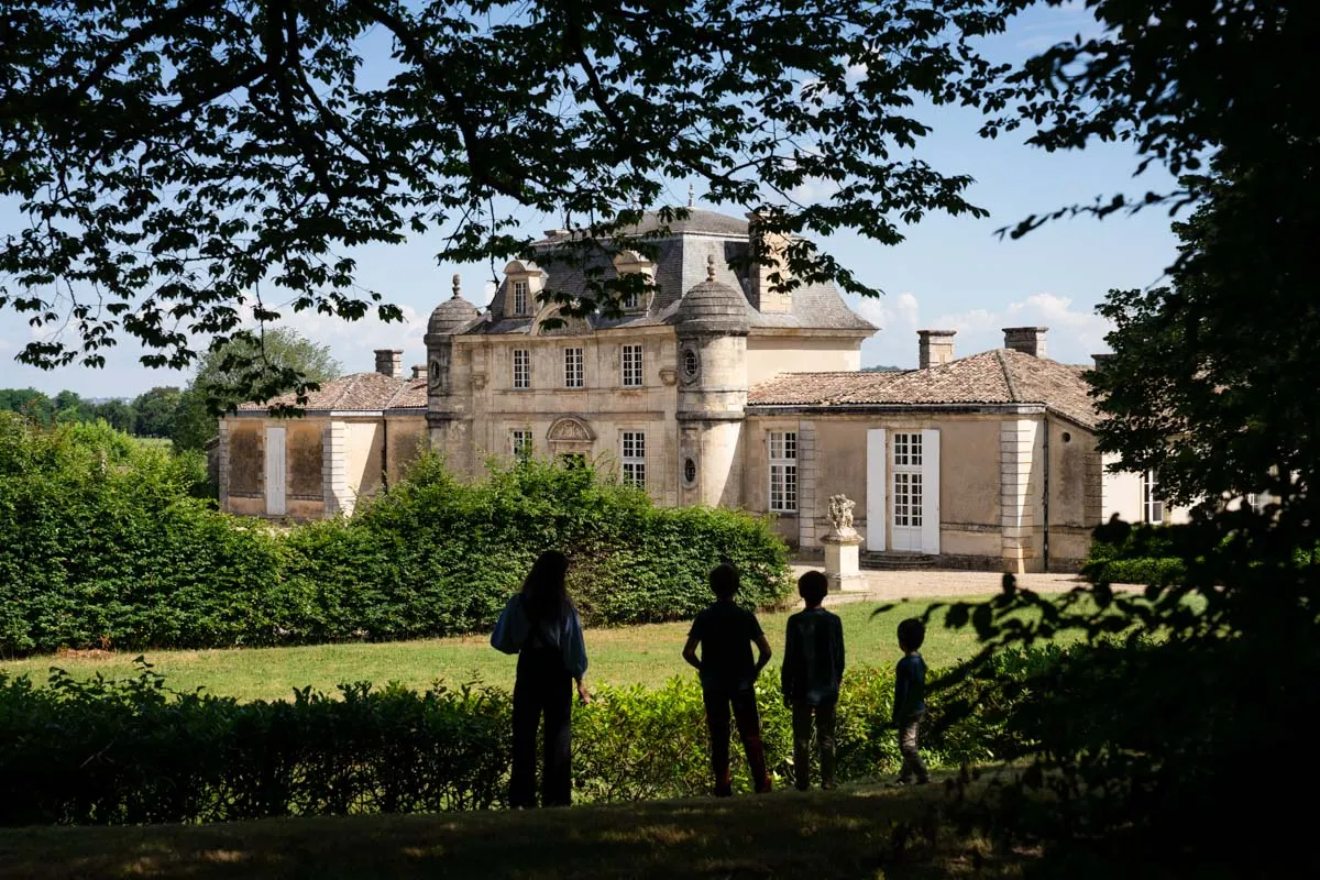Quatre visiteurs en tenue sombre debout dans un jardin face à un château du XVIIIe siècle de style néoclassique avec toits mansardés, architecture symétrique et haies taillées sous un ciel bleu.