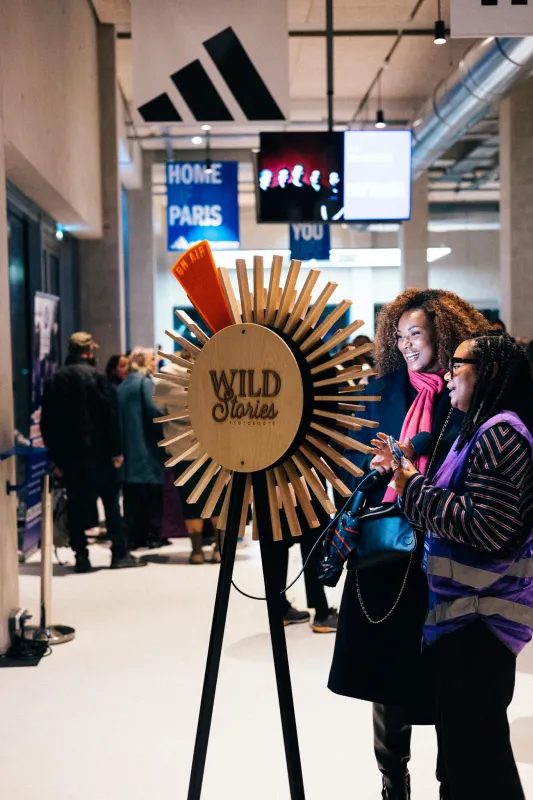 Deux visiteurs observent une grande installation interactive en forme de soleil doré portant le label 'Wild Stories' dans un espace moderne à Bordeaux, avec des écrans numériques visibles en arrière-plan.