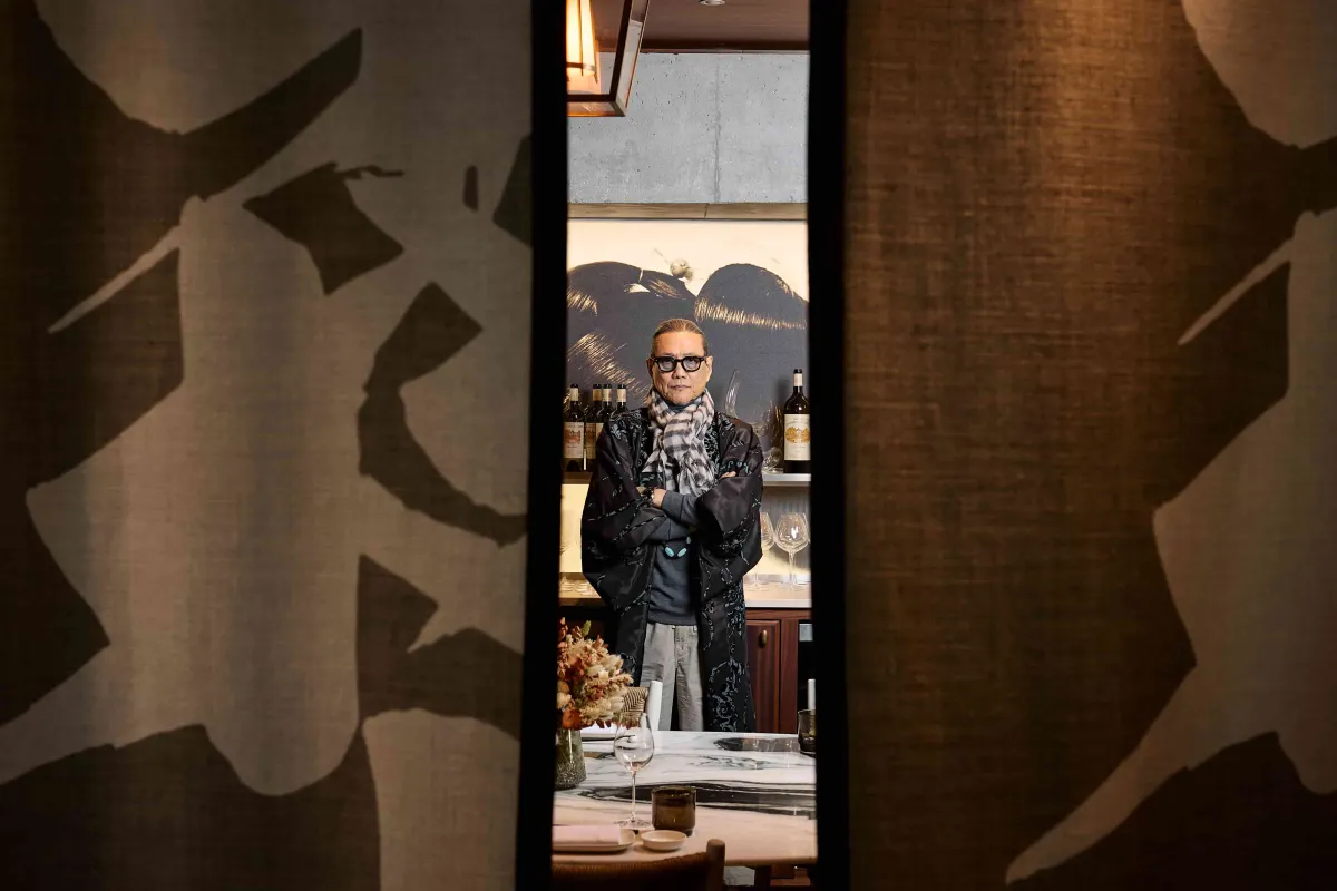Un homme portant des lunettes et une veste noire à motifs se tient dans un intérieur de restaurant élégant, encadré par une porte avec art mural décoratif, avec des bouteilles de vin et des éléments de salle à manger visibles en arrière-plan.