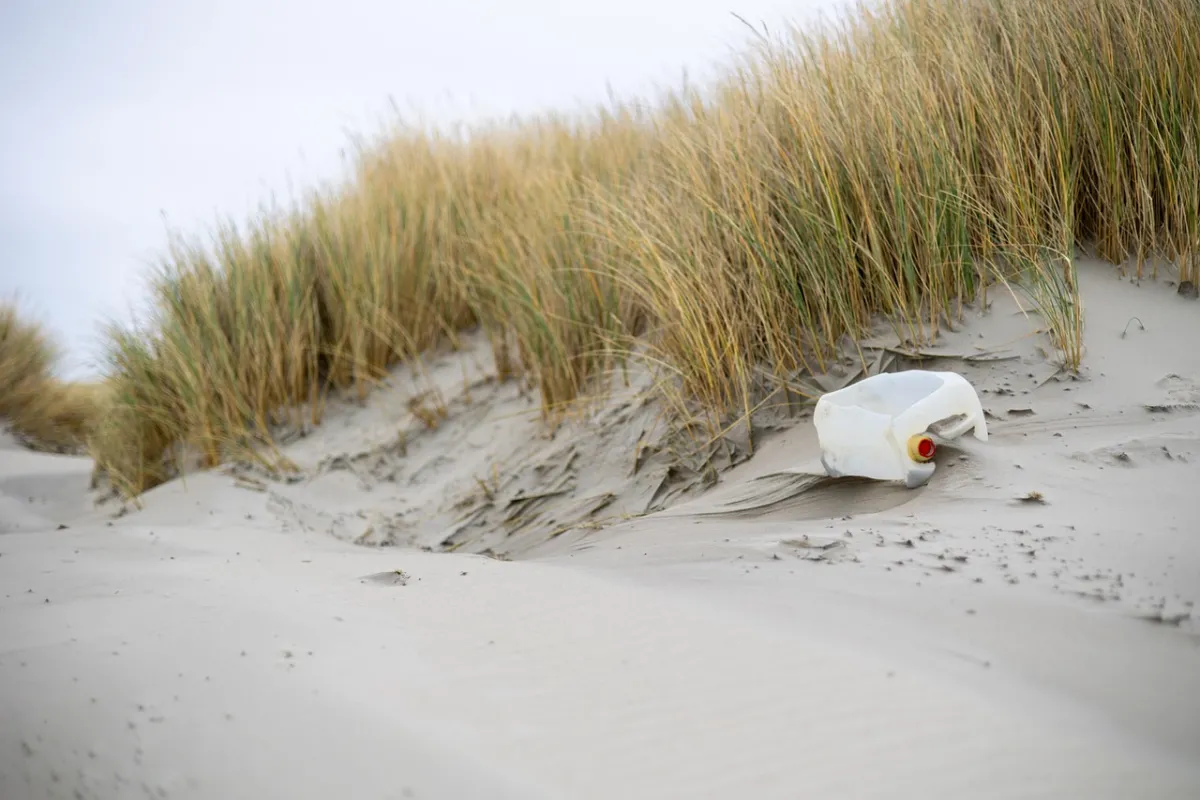 Bouteille en plastique blanche abandonnée sur une plage de sable avec des dunes d'oyat en arrière-plan, illustrant la pollution marine et la dégradation environnementale.