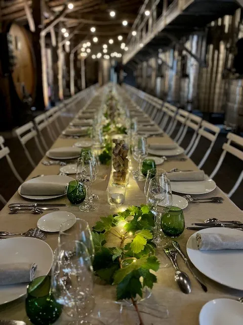 Table de banquet allongée dressée pour un dîner raffiné avec assiettes blanches, verres et un décor floral vert au centre, éclairée par des lumières suspendues dans une salle moderne.