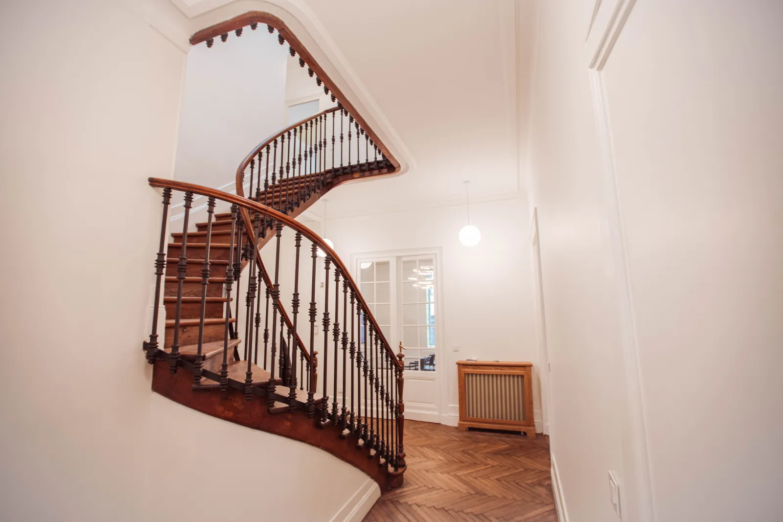 Escalier hélicoïdal élégant en bois avec rampes métalliques dans un couloir lumineux aux murs blancs et parquet chevrons, éclairé par un puits de lumière.
