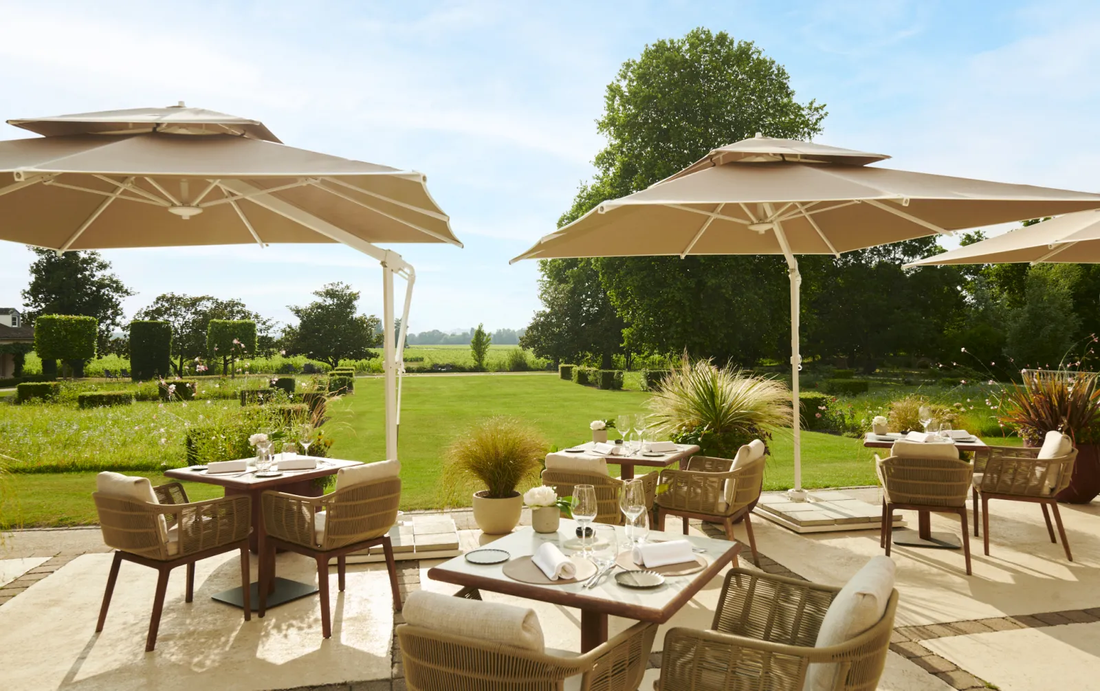 Terrasse élégante de restauration extérieure au Château Hôtel Grand Barrail avec parasols porte-à-faux beiges, mobilier en osier, couverts de table et jardins aménagés donnant sur des champs verdoyants près de Bordeaux.