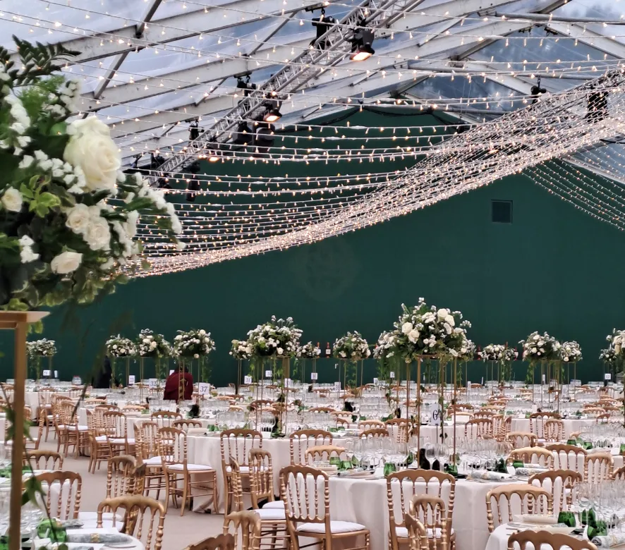 Salle de réception de mariage élégante avec tables rondes en lin blanc, chaises chiavari en bois, hauts centres de table fleuris blancs, mur vert en arrière-plan et guirlandes lumineuses décoratives au plafond.