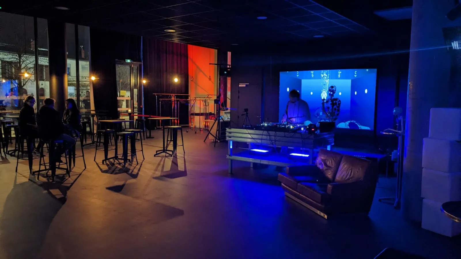 Espace événementiel moderne de L'Estrade à Bordeaux avec éclairage ambiant bleu, platine DJ sur scène, écran géant, sièges en cuir et visiteurs aux tables hautes près des baies vitrées la nuit.