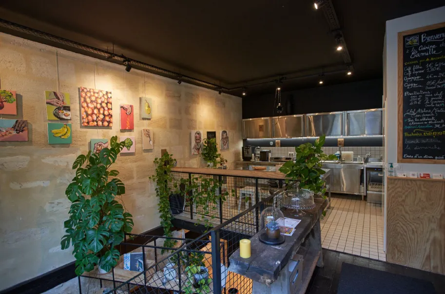 Intérieur d'une cuisine de restaurant moderne avec appareils en acier inoxydable, œuvres d'art culinaires décoratives sur les murs, plantes vertes, et une rambarde de comptoir en métal noir avec un menu ardoise visible à droite.