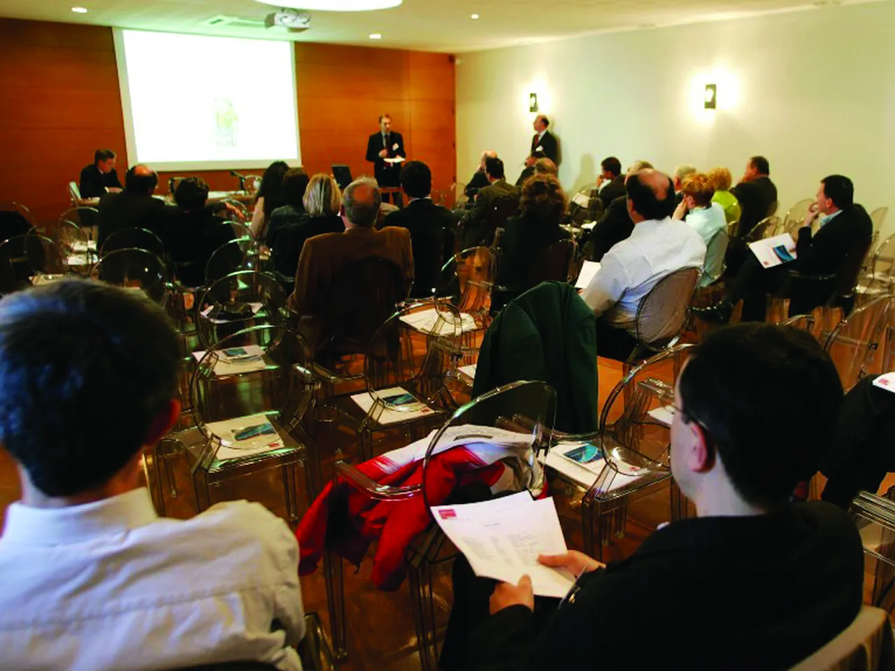 Salle de conférence professionnelle avec des participants assis en demi-cercle face à deux orateurs, un écran de projection affichant du contenu sur un mur orange.