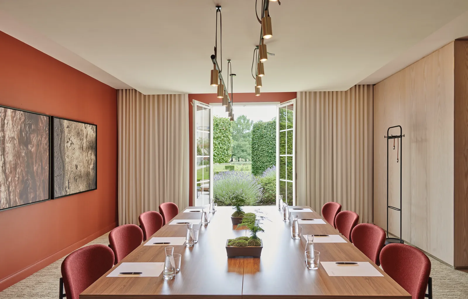 Salle de conférence contemporaine avec table en bois massif, chaises à dossier arrondi en velours bordeaux, mur d'accent orange, baies vitrées donnant sur des jardins paysagés avec lavande, luminaires suspendus et rideaux verticaux.