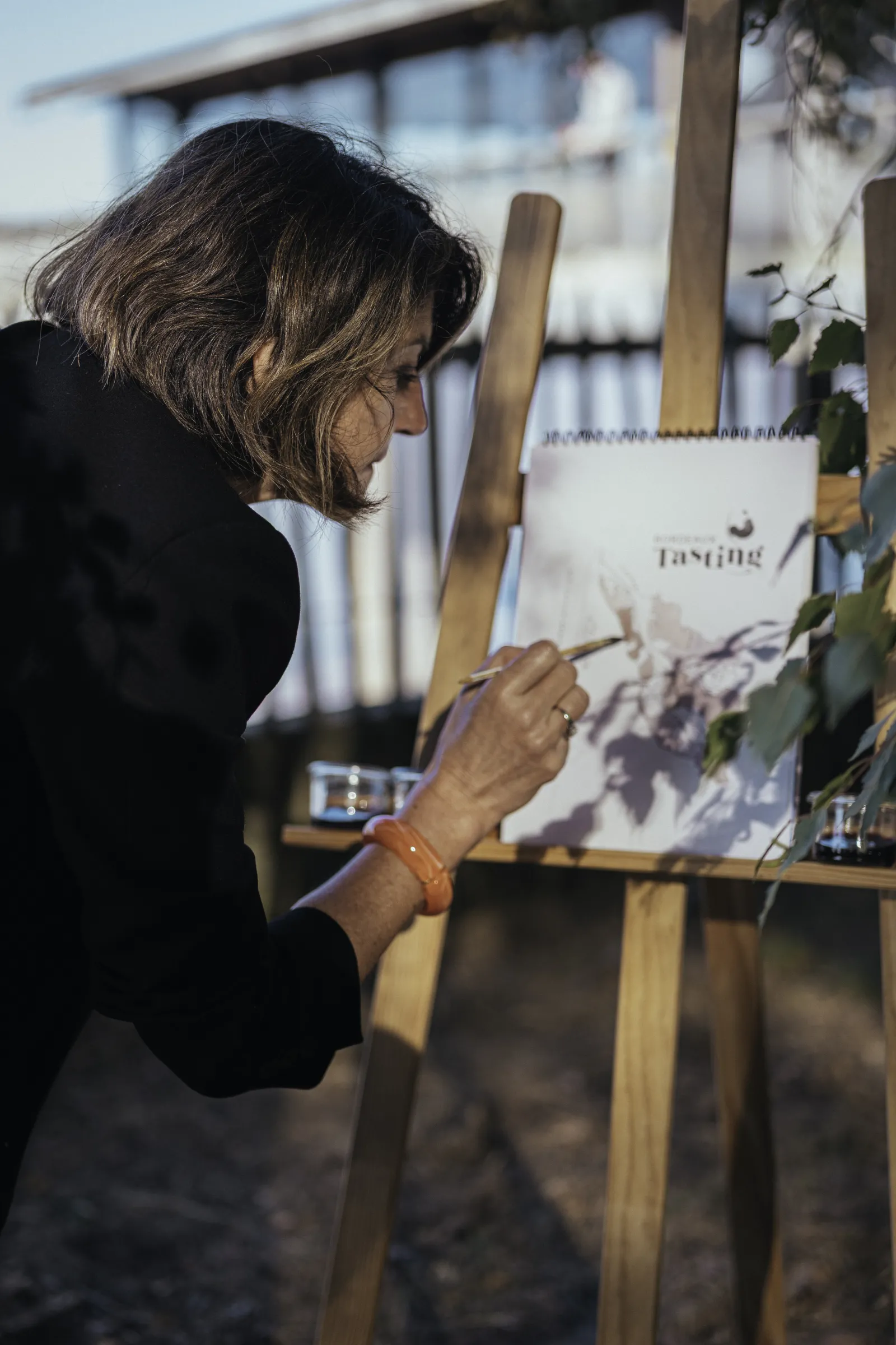 Femme en blazer noir écrivant sur une fiche de dégustation de vin lors d'un événement viticole en plein air à Bordeaux, avec chevalet en bois et bouteilles de vin visibles en arrière-plan.