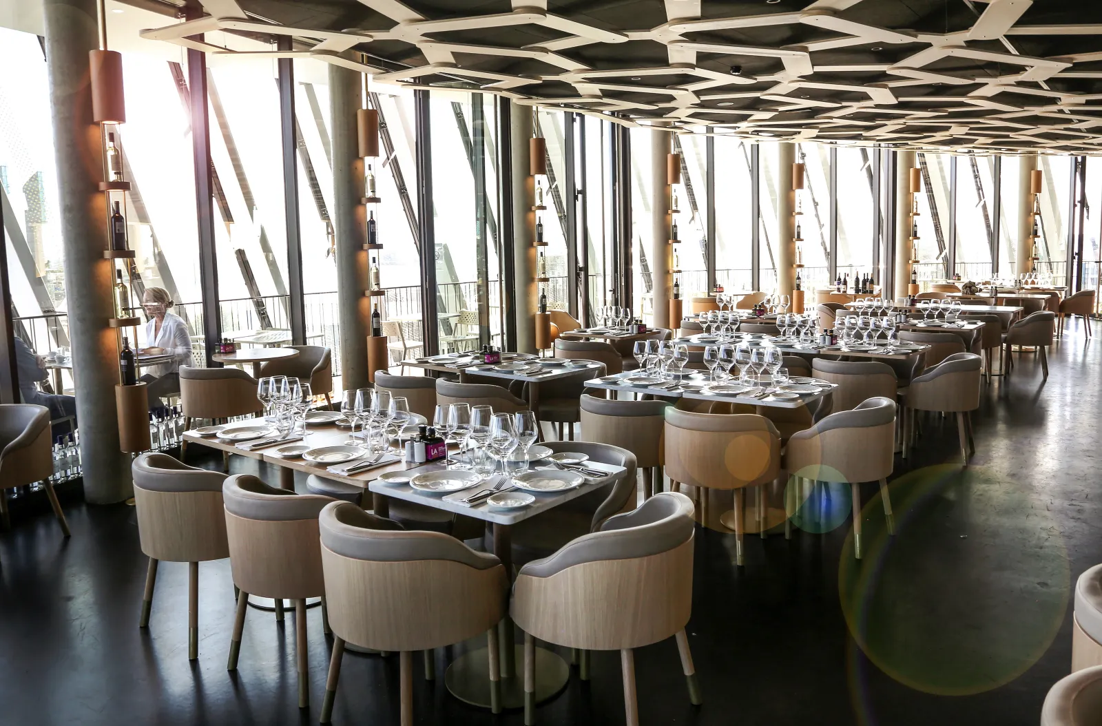 Restaurant gastronomique contemporain avec baies vitrées panoramiques surplombant Bordeaux, chaises rembourrées beige, tables noires dressées, plafond géométrique, et éléments sculpturaux élancés.