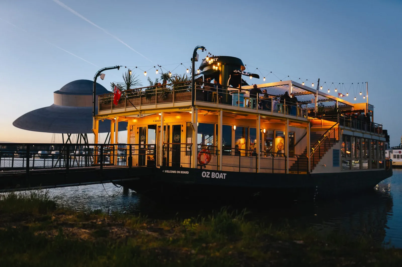 L'OZ Boat, un bateau de croisière fluviale jaune et noir amarré à Bordeaux avec pont illuminé au crépuscule, disposant d'espaces intérieurs et d'une terrasse d'observation supérieure.