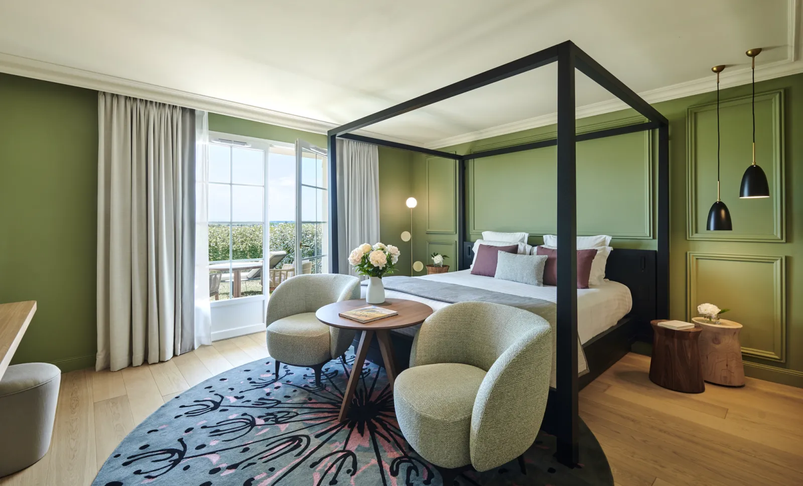 Suite luxueuse de l'hôtel château Grand Barrail à Bordeaux avec un lit à quatre colonnes noir, murs verts, fauteuils crème, table basse ronde en bois, grandes fenêtres donnant sur les jardins et éclairage suspendu élégant.