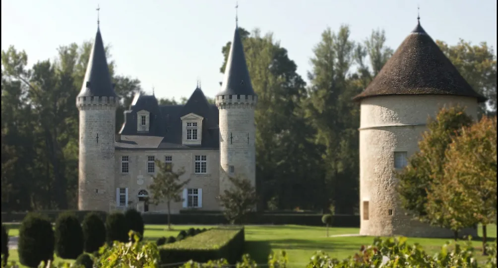 Château d'Agassac près de Bordeaux, présentant un château en pierre blanche avec deux tours élancées aux toits pointus noirs, un toit mansardé central et une tour ronde séparée à droite, entouré de jardins aménagés avec pelouse verte et topiaires.