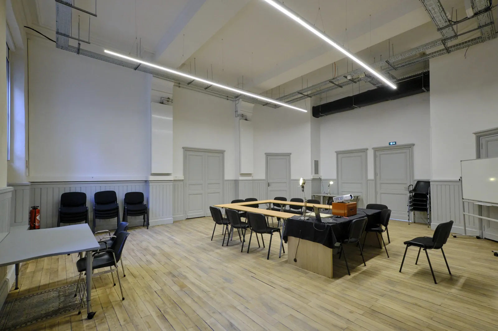 Salle de réunion moderne à La ManuCo à Bordeaux avec sol en bois, table longue en bois, chaises noires, murs à lambris blanc, éclairage linéaire au plafond et plusieurs portes.