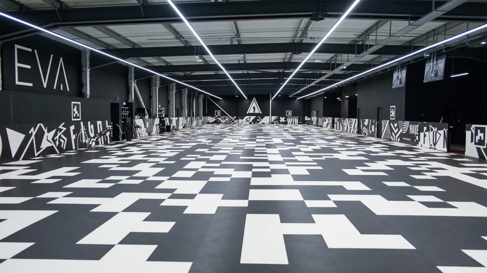 Vaste espace intérieur moderne avec sol à motif géométrique noir et blanc, éclairage linéaire LED au plafond, et signalisation néon blanche sur murs sombres au lieu EVA Bordeaux Bègles.