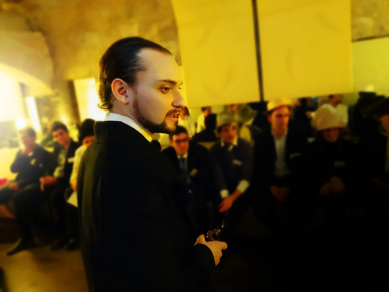 Homme aux cheveux noirs et bouc vêtu d'une veste noire formelle, s'adressant à une foule dans un lieu intérieur aux murs jaunes à Bordeaux