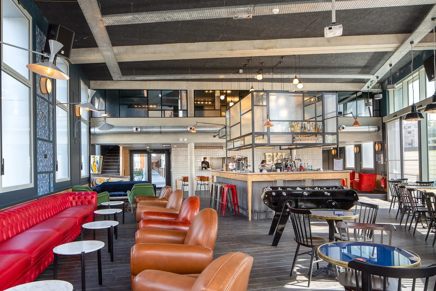 Hall d'accueil et espace lounge moderne de l'Eklo Bordeaux avec sièges en cuir rouge et caramel, bar central en bois, baby-foot, plafond en béton apparent au design industriel et grandes baies vitrées.