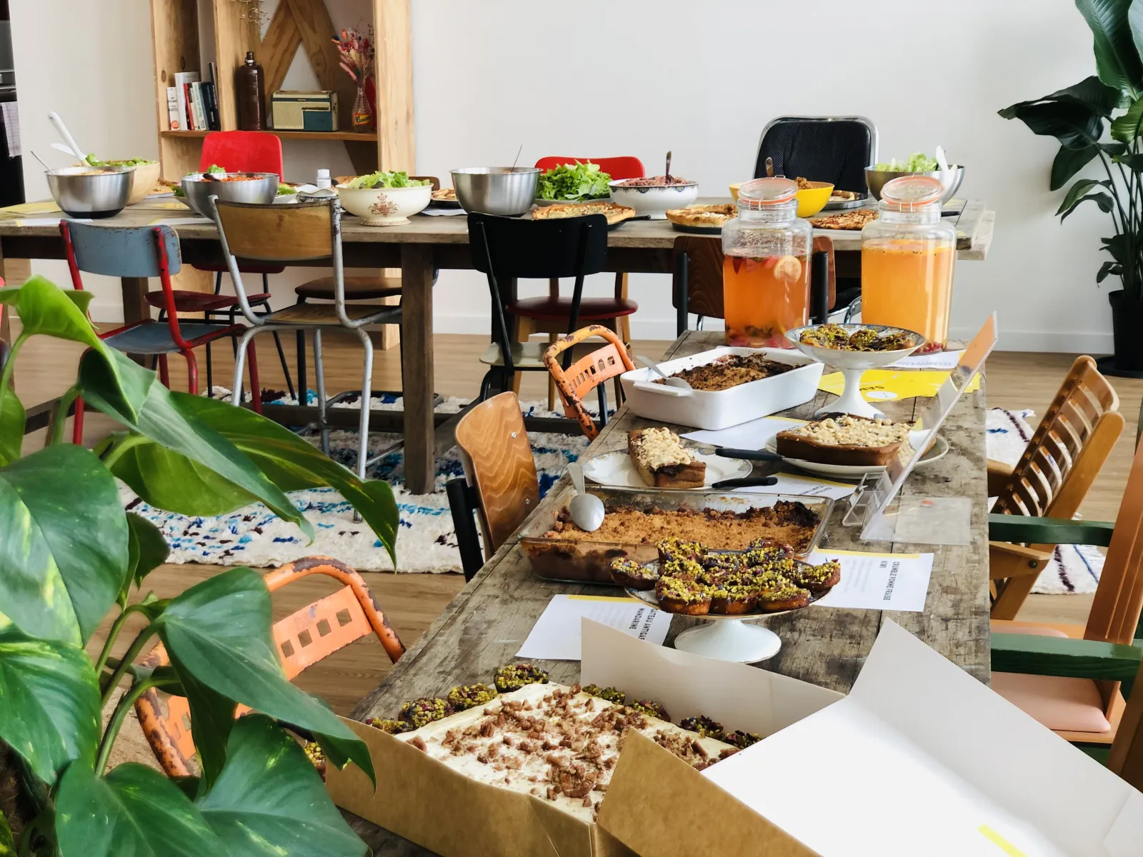 Un repas communautaire à Bordeaux présentant plusieurs tables chargées de plats faits maison, pâtisseries, salades fraîches et boissons en bocaux, avec des chaises variées et des plantes en pot créant une atmosphère de convivialité inclusive.